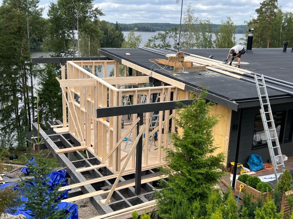Puurunkoinen laajennus rakenteilla tumman talon kylkeen järven rannalla
