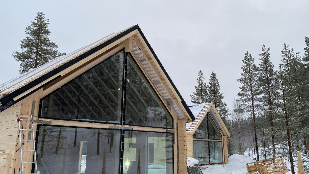 Rakenteilla oleva moderni puutalo, jossa lasinen päätyseinä, lumisessa metsämaisemassa