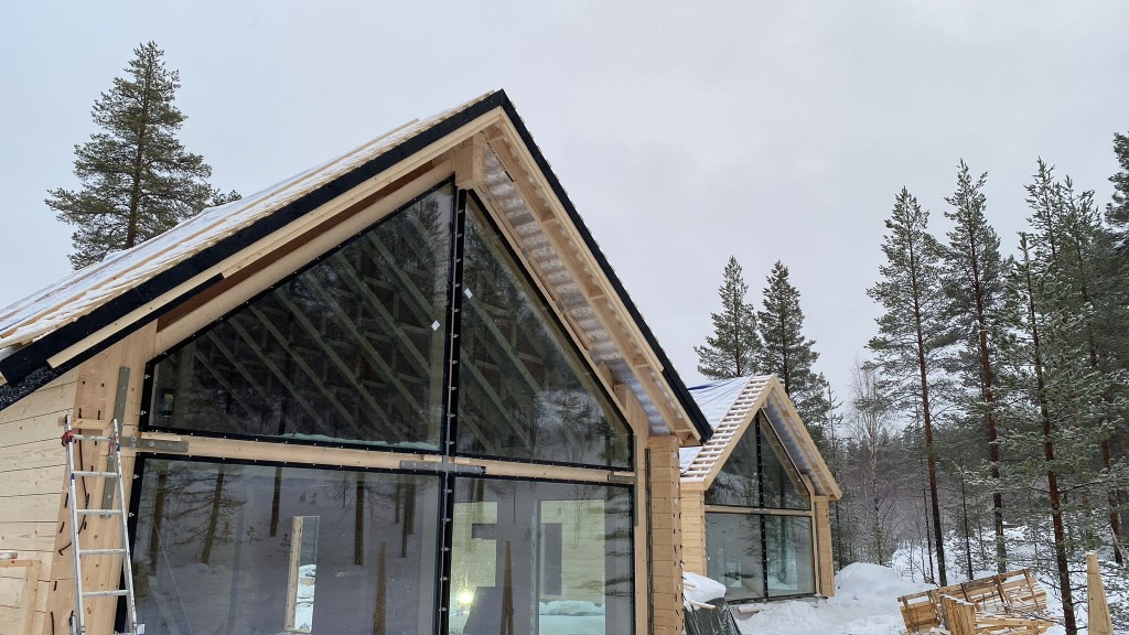 Rakenteilla oleva moderni puutalo, jossa lasinen päätyseinä, lumisessa metsämaisemassa